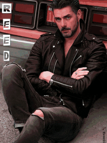 a man in a leather jacket sits on the ground in front of a red truck