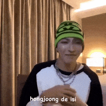a man wearing a green beanie and a necklace is sitting in a room with a curtain behind him .