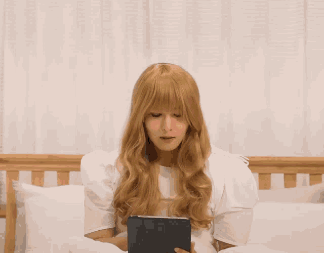 a woman with long blonde hair is sitting on a bed holding a tablet