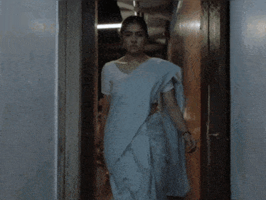 a woman in a white shirt and blue saree walks down a hallway