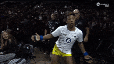 a woman in a white ufc shirt and yellow shorts is dancing in front of a crowd