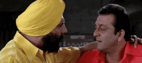 a man wearing a yellow turban and a red shirt talks to another man