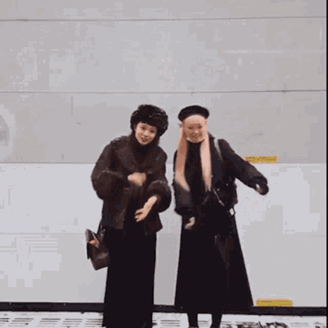 two women are standing next to each other in front of a wall wearing black clothes .
