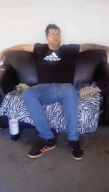 a man wearing an adidas shirt sits on a couch with his arms outstretched