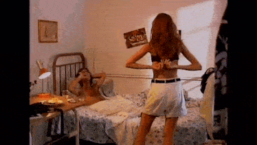 a woman in a bra is standing in a bedroom next to a man in a bed .