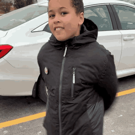 a young boy in a black jacket stands in front of a car