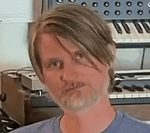 a man with long hair and a beard is standing in front of a keyboard in a room .