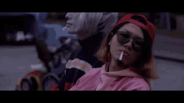 a woman in sunglasses is smoking a cigarette next to a man in a baseball cap .