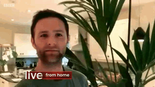 a man stands in front of a plant with the words live from home behind him