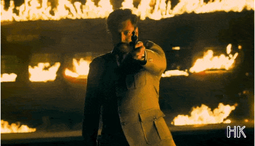 a man in a suit is pointing a gun in front of a burning building with the letters hk below him