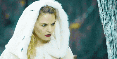 a woman in a white fur coat is standing next to a tree .