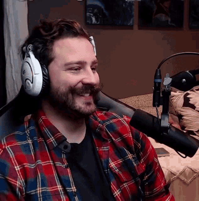 a man wearing headphones and a plaid shirt is sitting in front of a microphone and smiling