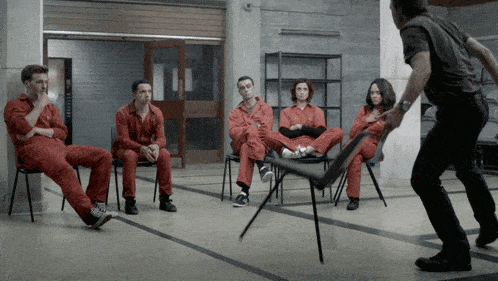 a group of people in red jumpsuits sit in chairs while a man throws a chair at them