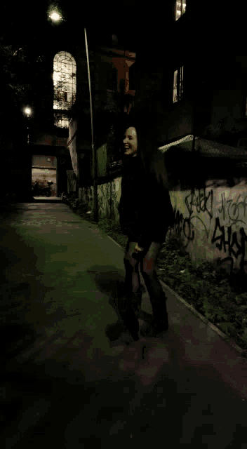 a woman is walking down a street at night with graffiti on the wall behind her