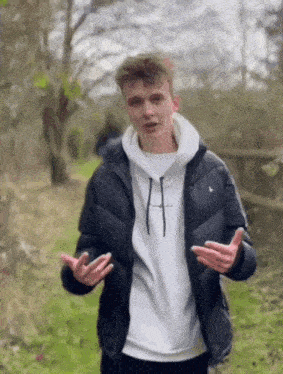 a young man wearing a black jacket and a white sweatshirt is standing in a field with his hands outstretched .