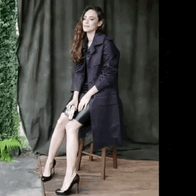 a woman in a trench coat is sitting on a wooden stool