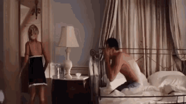 a man without a shirt is sitting on a bed with a woman standing next to him .