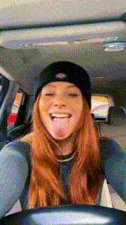 a woman with red hair wearing a black beanie is sticking her tongue out in a car .