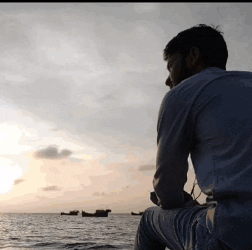 a man sits on the edge of a body of water with boats in the distance