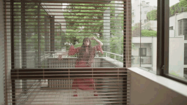 a woman in a red dress is standing in front of a window with blinds on it
