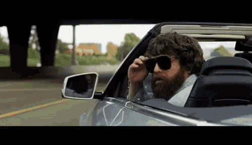 a bearded man wearing sunglasses is driving a convertible car