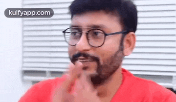 a man with a beard and glasses is sitting in front of a window with blinds .