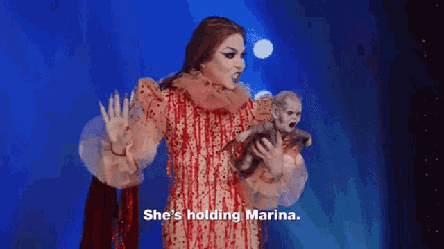 a drag queen in a red dress is holding a monkey and saying `` she 's holding marina . ''