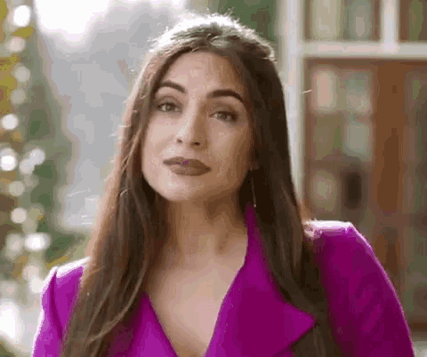 a woman in a purple jacket is standing in front of a window and looking at the camera .
