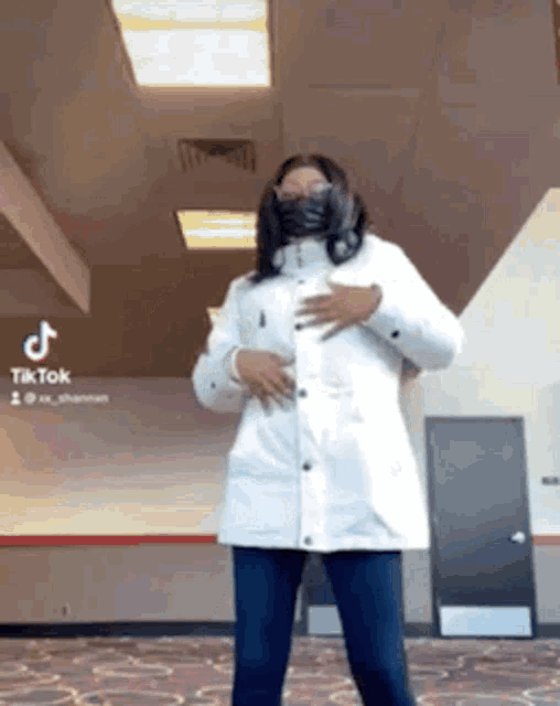 a woman wearing a white coat and a black mask is standing in a room .