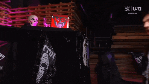 a wrestler with long hair and tattoos is standing in front of a skull and a box .