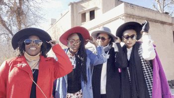 four women wearing hats and sunglasses pose for a picture in front of a building