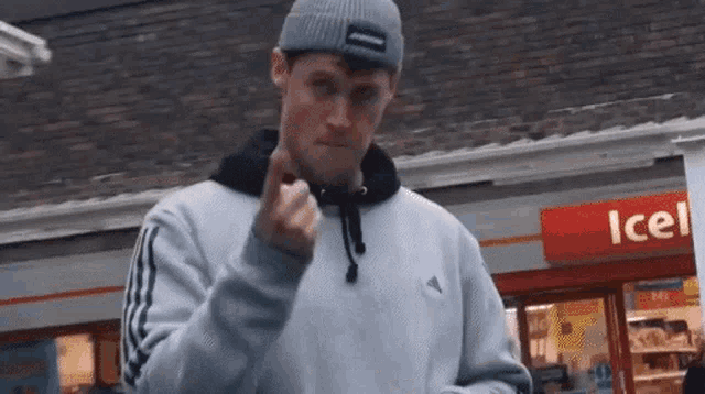 a man wearing a beanie and a sweatshirt is giving the middle finger in front of a store .