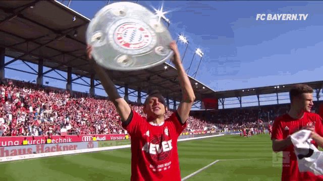 a soccer player holds up a trophy in front of a crowd with fc bayern.tv written on the bottom