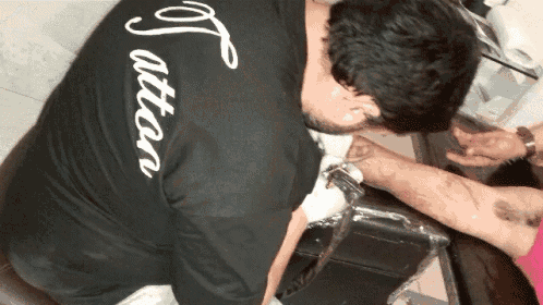 a man is getting a tattoo on his arm while wearing a black shirt that says " attare " on the back