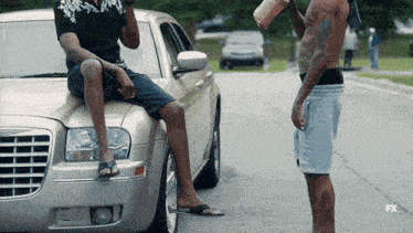 a man sits on the hood of a car while another man stands nearby