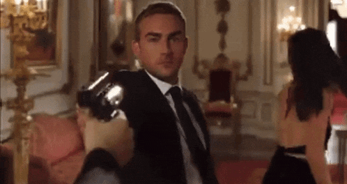 a man in a suit and tie is holding a gun in a room with a woman in a dress behind him .