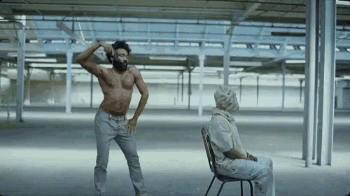 a man without a shirt is dancing in an empty room while another man sits in a chair