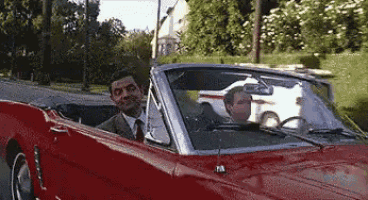 a man in a suit and tie is driving a red convertible car on a street .