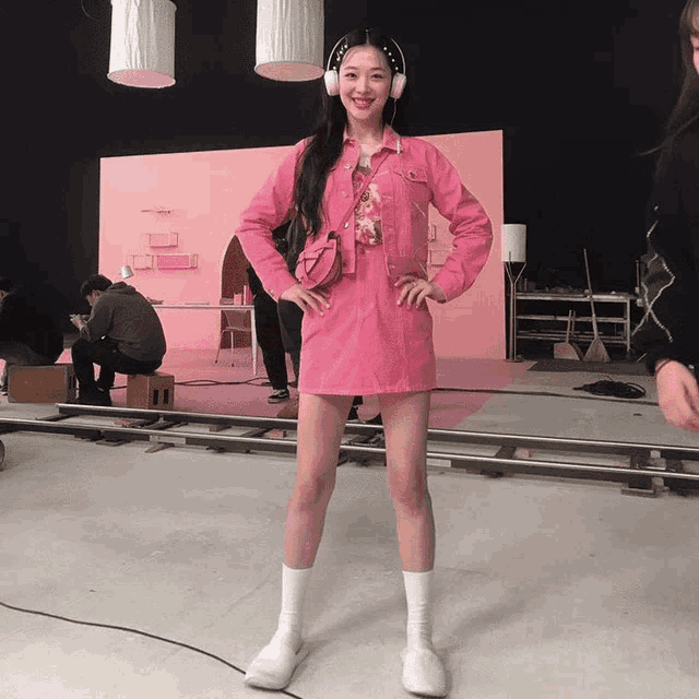 a woman in a pink dress and white socks stands on a set with her hands on her hips