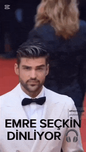 a man in a white suit and black bow tie is walking down a red carpet .