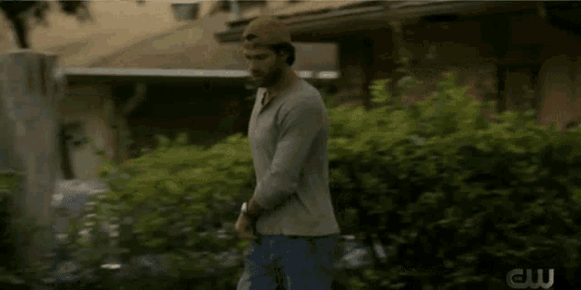 a man wearing a baseball cap and a grey shirt is walking in front of a house with a cw logo behind him