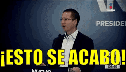 a man in a suit stands in front of a microphone with the words " esto se acaba " above him