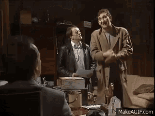 a group of men are standing around a table in a living room talking to each other .