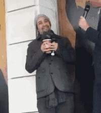 a man in a suit is holding a cup of coffee while being interviewed by a man holding a microphone .