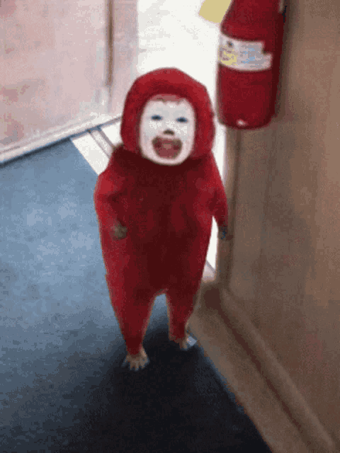 a baby in a red costume with a white face is standing in a hallway next to a red fire extinguisher