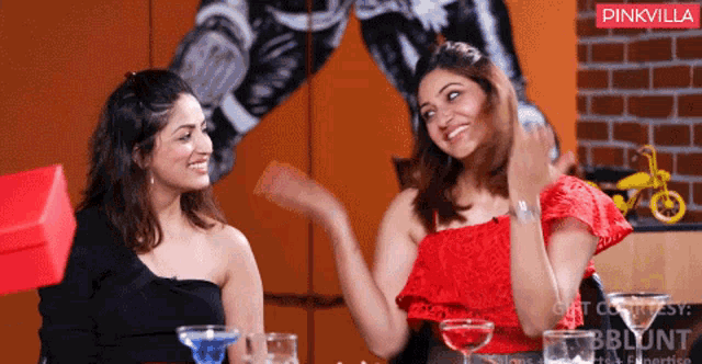 two women are sitting at a table with drinks in front of a wall with a pinkvilla logo