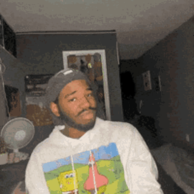 a man wearing a spongebob sweatshirt and a hat is sitting in a room