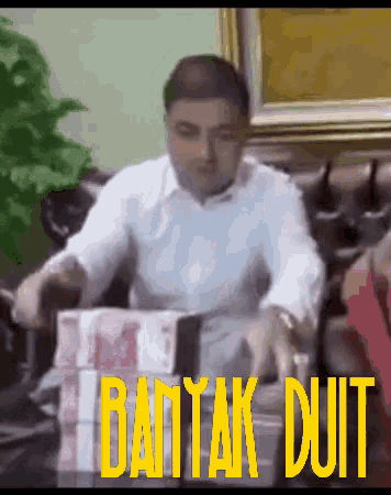 a man is sitting on a couch with a stack of money in front of him that says bantak duit on it
