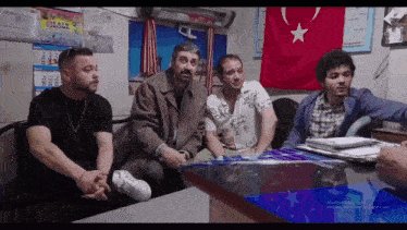 a group of men sit around a table in front of a flag