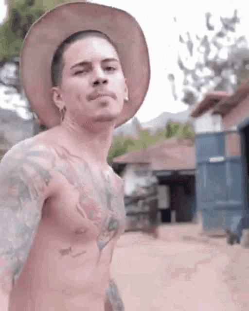 a shirtless man wearing a cowboy hat is standing in front of a house .